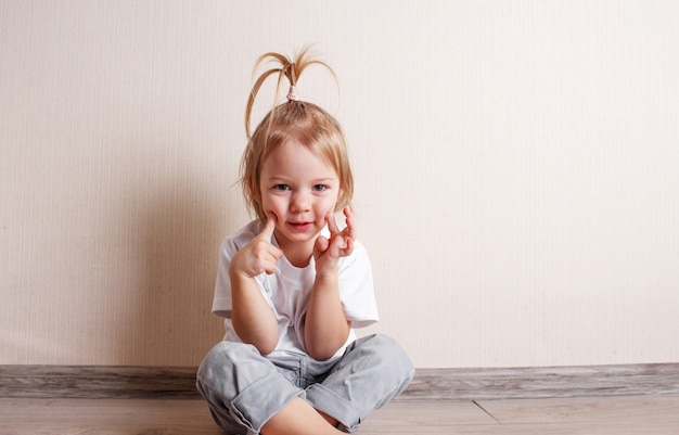 A little girl in a white T-shirt sits on the floor of the room against the wall.