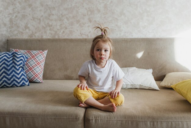 흰색 티셔츠와 노란색 바지를 입은 어린 소녀가 사려 깊은 표정으로 집에서 소파에 앉아 있습니다.