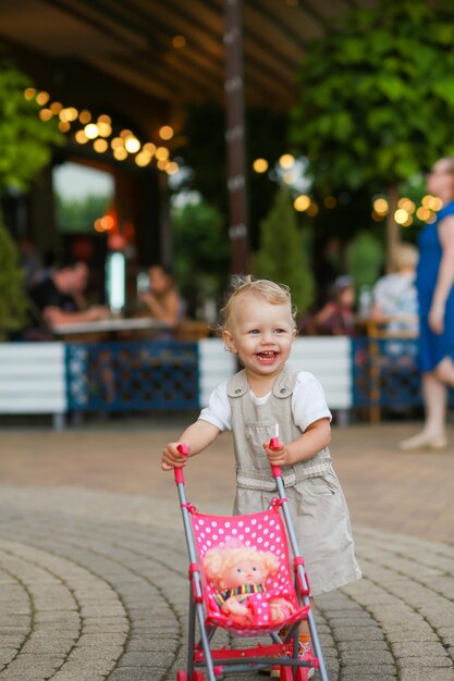 A little girl who has recently learned to walk is pushing a small toy stroller against a blurred bac