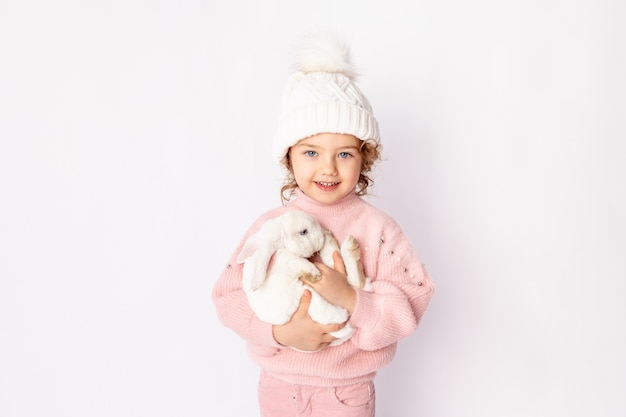 A little girl in winter clothes holds a rabbit on a white background.