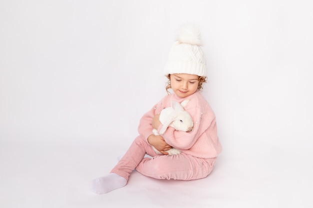 A little girl in winter clothes holds a rabbit on a white background.
