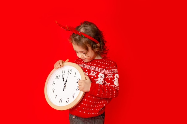 A little girl in winter clothes holds a watch on a red background. New year's concept, space for text