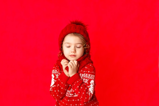 A little girl in winter clothes makes a wish with her eyes closed on a red background. New year's concept, , place for text