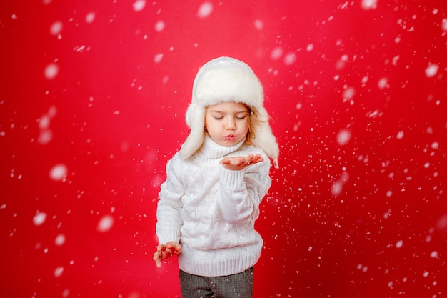 Little girl in winter hat blowing on snow red background