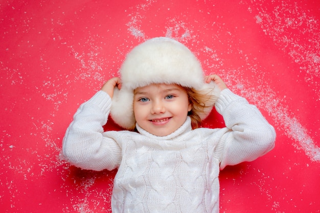 a little girl in a winter hat is lying on the floor in the snow on a red background