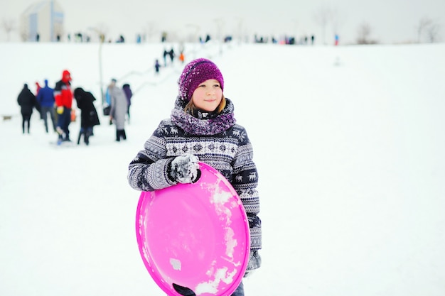 スカンジナビアのプリントが施された冬のアウターの少女は、雪の斜面に対して氷のそりと笑顔を保持しています。冬のエンターテイメント