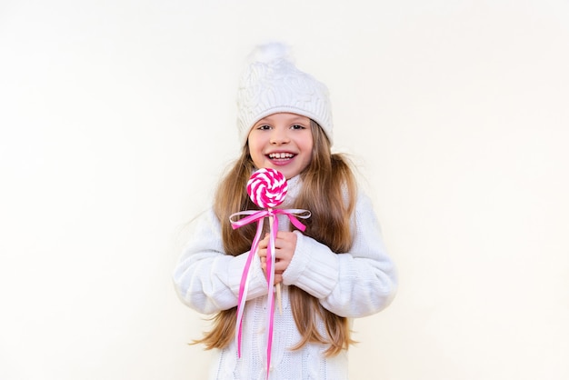 A little girl in a winter sweater and hat holds a delicious lollipop and smiles on a white isolated background.