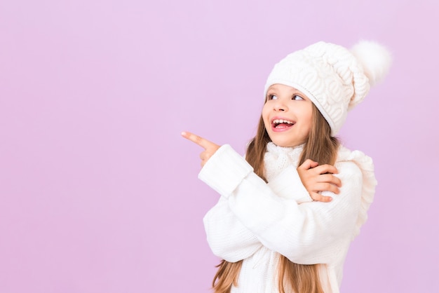 Маленькая девочка в зимнем свитере и шляпе указывает на вашу рекламу на изолированном розовом фоне.