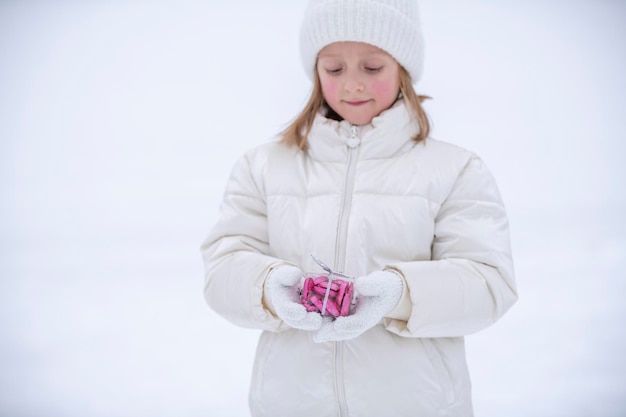 A little girl in winter white clothes in the snow holding a transparent box with candies in the shape of hearts