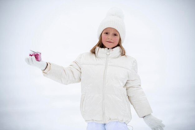 Una bambina in abiti bianchi invernali nella neve con in mano una scatola trasparente con caramelle a forma di cuore