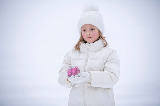 Una bambina in abiti bianchi invernali nella neve con in mano una scatola trasparente con caramelle a forma di cuore