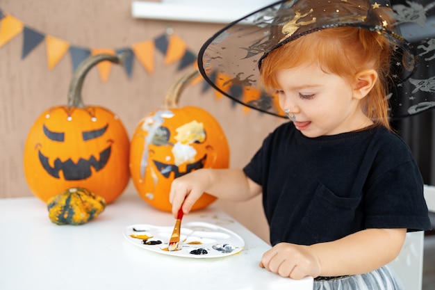 A little girl in a witch costume paints pumpkins for halloween at home the concept of childrens crea...