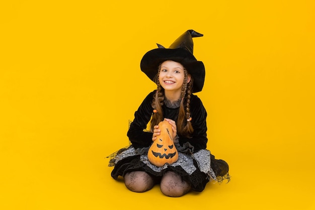 A little girl in a witch costume and a pointed hat is sitting in full height legs tucked under her and holding a pumpkin in her hands child is happy with halloween yellow isolated background