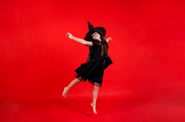 A little girl in a witch costume and with a hat jumps on a red background with a copy of the space