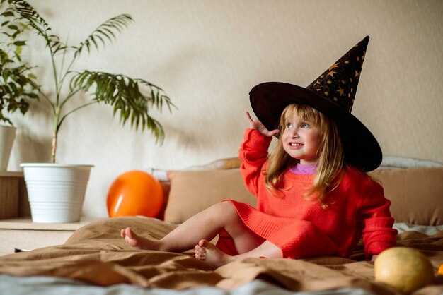 Little girl in a witch hat with an orange ball in her hand. halloween concept.active games at home. laughs emotionally and jumps on the bed.