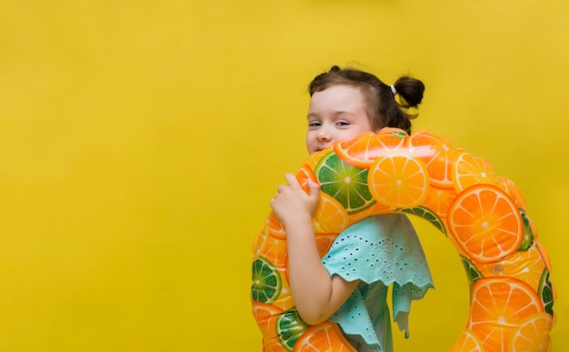 Фото Маленькая девочка с надувным кругом с апельсинами стоит боком на желтом фоне с копией пространства