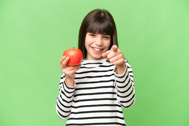 격리된 크로마 키 배경 위에 사과를 들고 있는 어린 소녀가 자신감 있는 표정으로 당신을 손가락질합니다