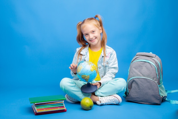 A little girl with a backpack and books is sitting on a blue background