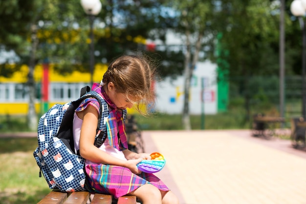 Little girl with a backpack and in a school uniform in the\
school yard plays pop it toy. back to school, september 1. the\
pupil relaxes after lessons. primary education, elementary class\
for student