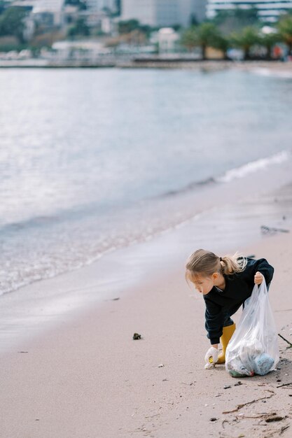 Little girl with a bag in gloves picks up garbage from a sandy beach near the sea