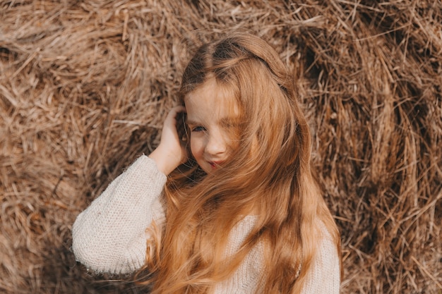 A little girl with beautiful blond hair sits on a field near a roll of hay. portrait of a child in a knitted hat.