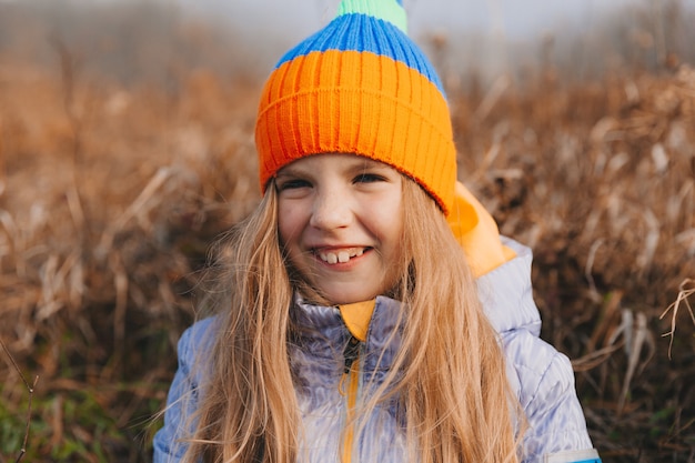 A little girl with beautiful blond hair sits on a field near a roll of hay. portrait of a child in a knitted hat.