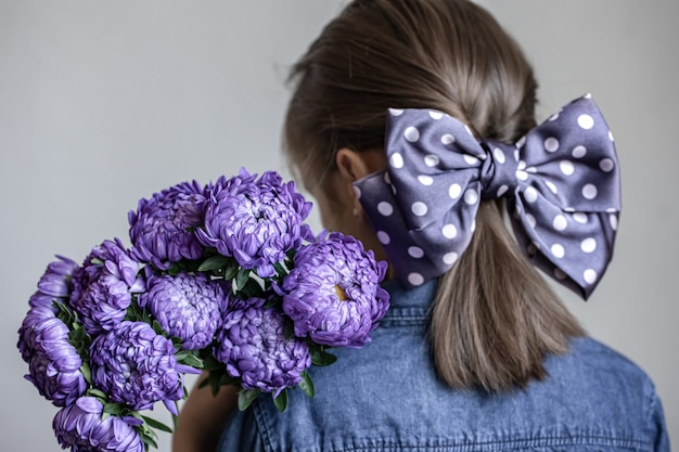 Little girl with a beautiful bow on her hair holds a bouquet of blue chrysanthemums, back view.