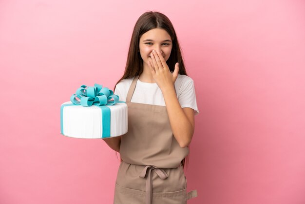 Bambina con una grande torta su sfondo rosa isolato felice e sorridente che copre la bocca con la mano