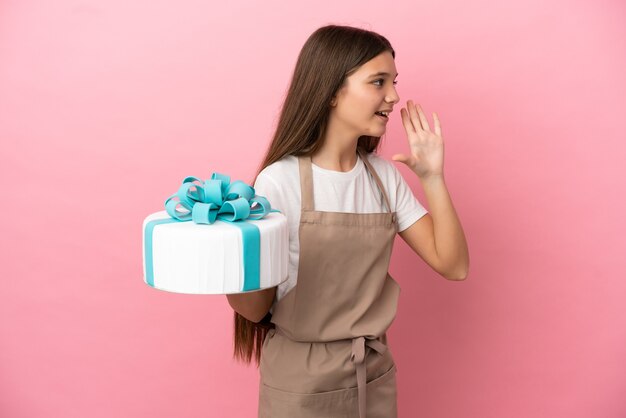 Little girl with a big cake over isolated pink background shouting with mouth wide open to the side