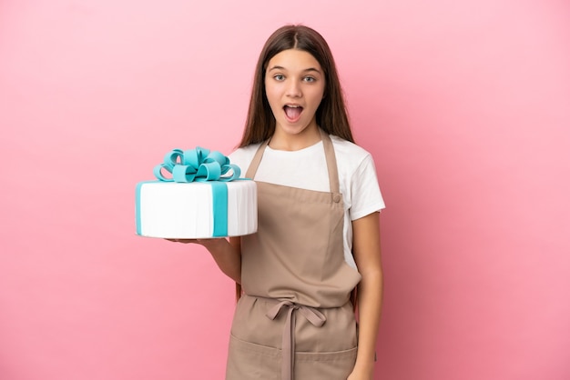 Bambina con una grande torta su sfondo rosa isolato con espressione facciale a sorpresa