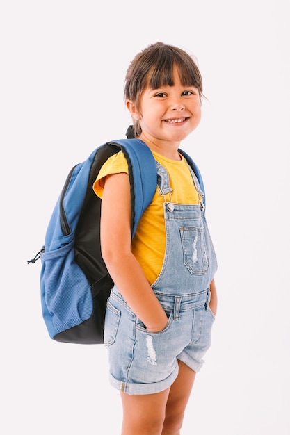 デニムのオーバーオールと青いTシャツを着た黒い髪の少女、白い背景の上に、彼女の側で、学校に戻る準備ができているバックパックを持っています