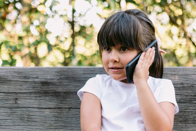 Маленькая девочка с черными волосами, одетая в белую футболку, сидит на скамейке в парке, разговаривает по мобильному телефону, с серьезным жестом.