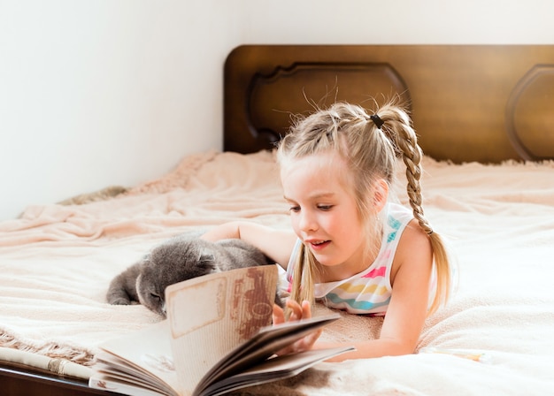 A little girl with blond hair lies on a bed at home with a cat reading a book. Stay at home. Home education for children.