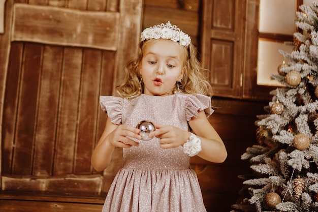 Photo little girl with blond hair in pink dress in new year background. girl blue eyes near christmas tree with ball in her hands.