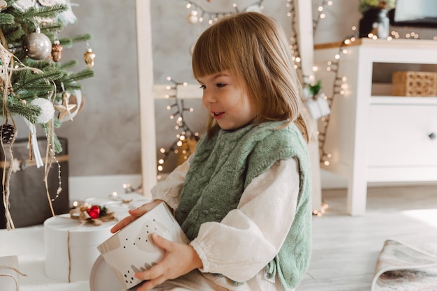 Little girl with blond hair sits on the floor near the Christmas tree with gifts.