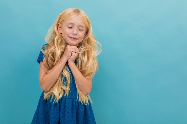 Little girl with blonde hair in blue dress is relaxed and enjoying, portrait isolated on blue