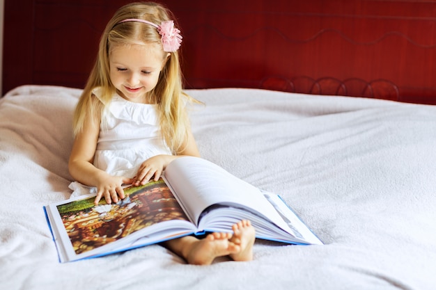 Little girl with blonde hair reading book on the bed