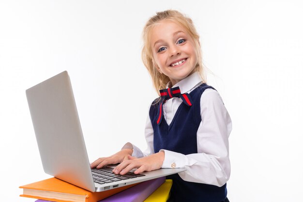 A little girl with blonde hair stuffed in a horse tail, large blue eyes and a cute face with a laptop on the books.