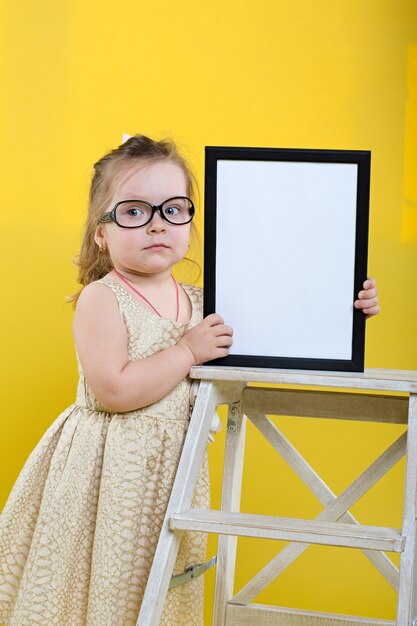 Photo little girl with board in beautiful dress and glasses on yellow background