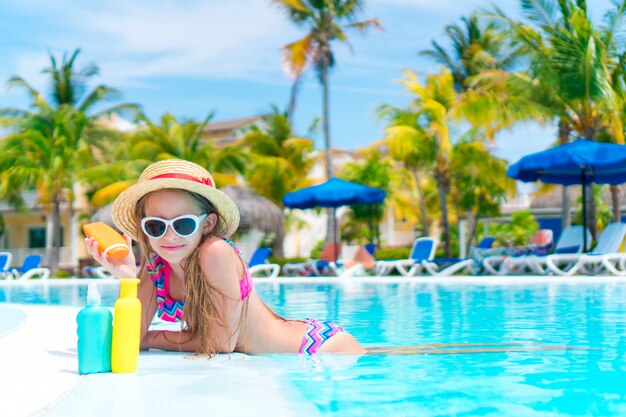Little girl with bottle of sun cream in swimming pool