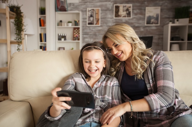 Photo little girl with braces and her mother having fun relaxing sitting on the couch using smartphone in living room.