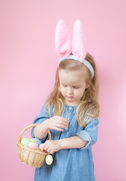 Little girl with bunny ears standing with wooden basket full of colorful Easter eggs