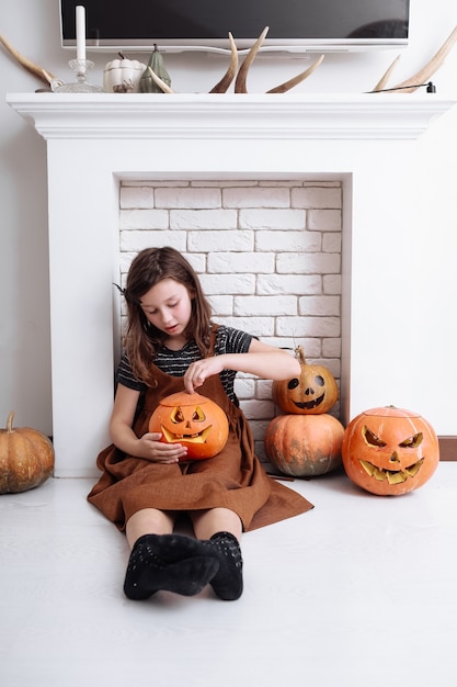 Little girl with carved pumpkin on Halloween at home sitting next to fireplace