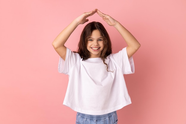 Bambina con un sorriso affascinante che si tiene per mano nel gesto del tetto della casa sopra la testa e sorridente