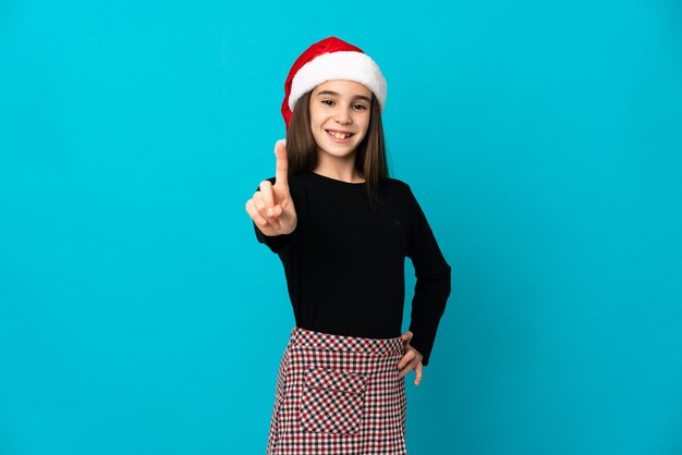 Little girl with Christmas hat isolated on blue background showing and lifting a finger