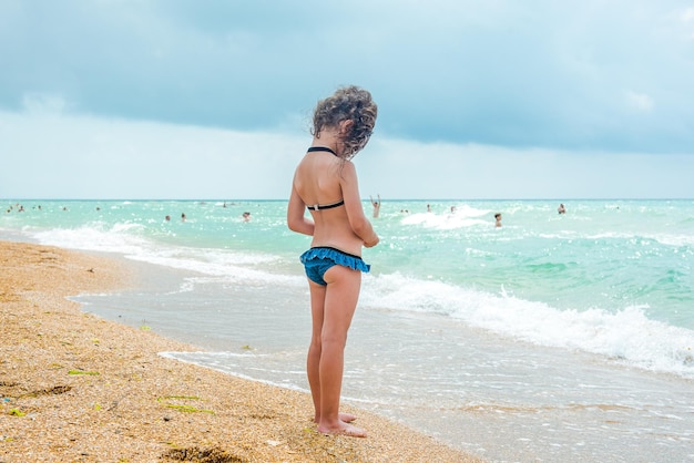 A little girl with curly hair in a blue bathing suit stands with her back to the camera on a sandy