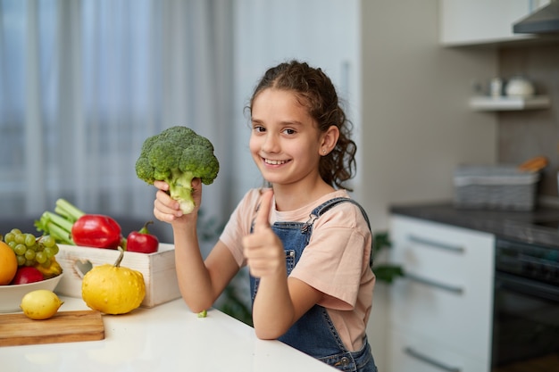 Little girl with curly hair, holding a broccoli and looking at camera, showing a thumbs, seated at table in the kitchen.