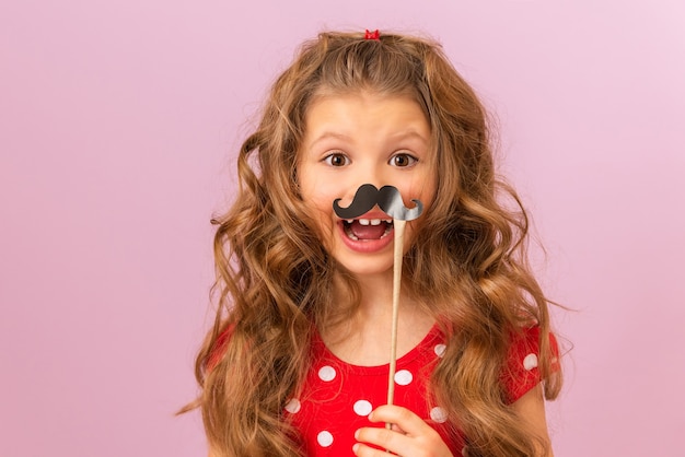 A little girl with curly hair holds a fancy paper moustache.