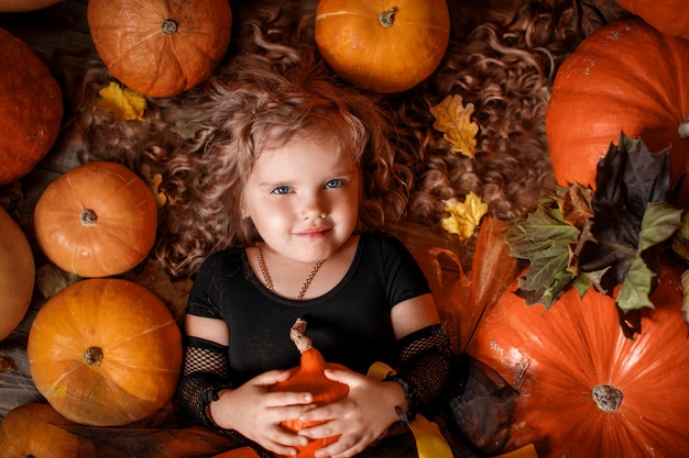 Little girl with curly hair in a witch costume lies among pumpkins