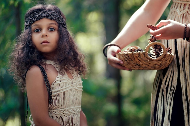 Foto una bambina con i capelli ricci scuri vestita da nativa nella foresta raccoglie dossi con sua madre